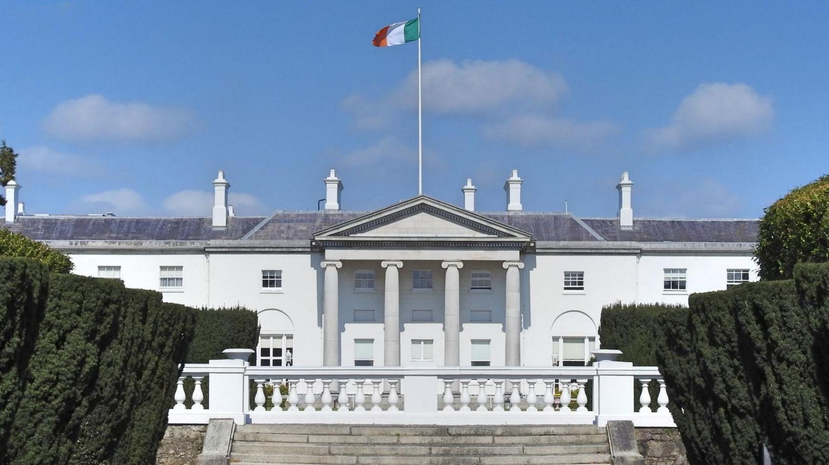 The exterior of Áras an Uachtaráin - the official residence of the Irish president. It is a white building, not dissimilar to the White House in Washington. It features four large columns at the front of it. An Irish tricolour is flown from a flagpole on the roof of the building.