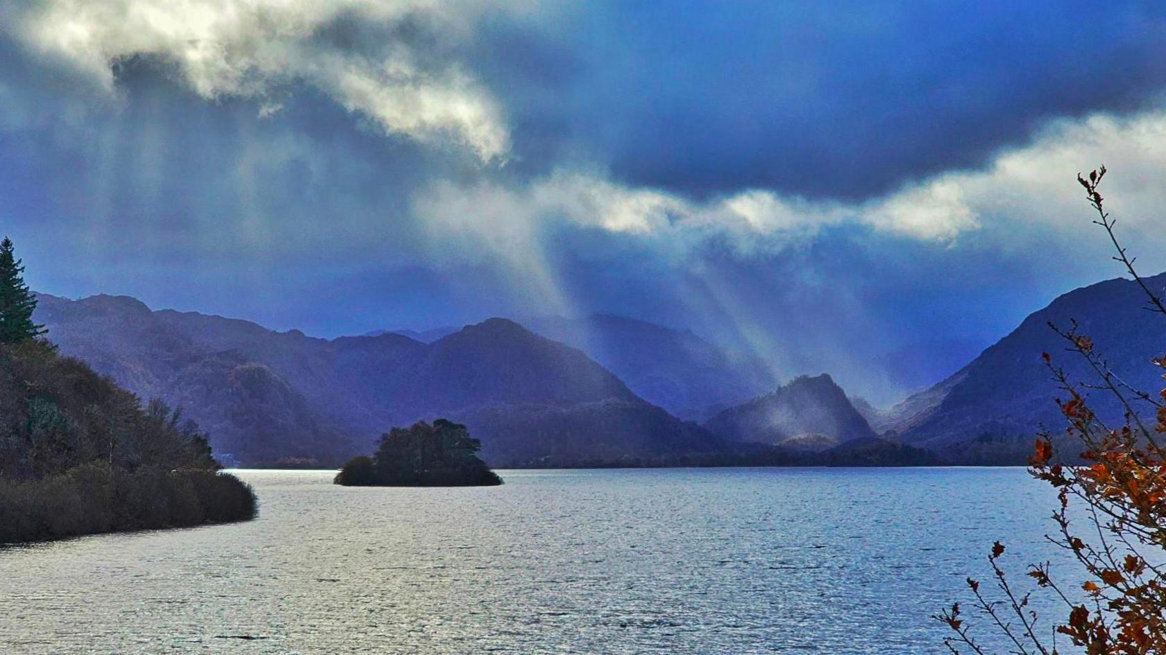 The water is very calm and of different shades of blue. There is white haze in front the hills beneath a backdrop of a blue and white cloudy sky. 