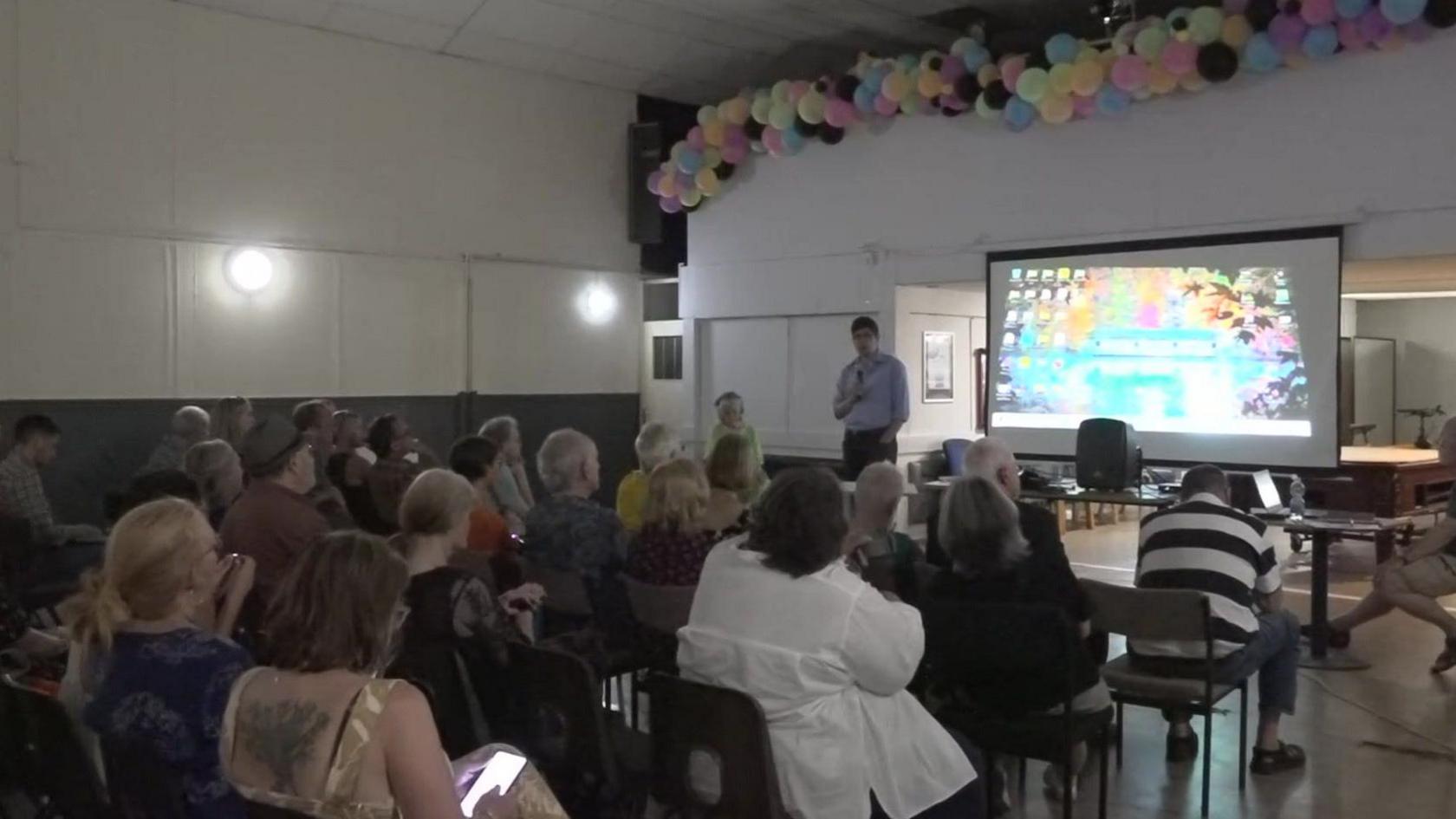 Cynulleidfa yn edrych ar sgrin lle mae Huw Thomas yn siarad 
