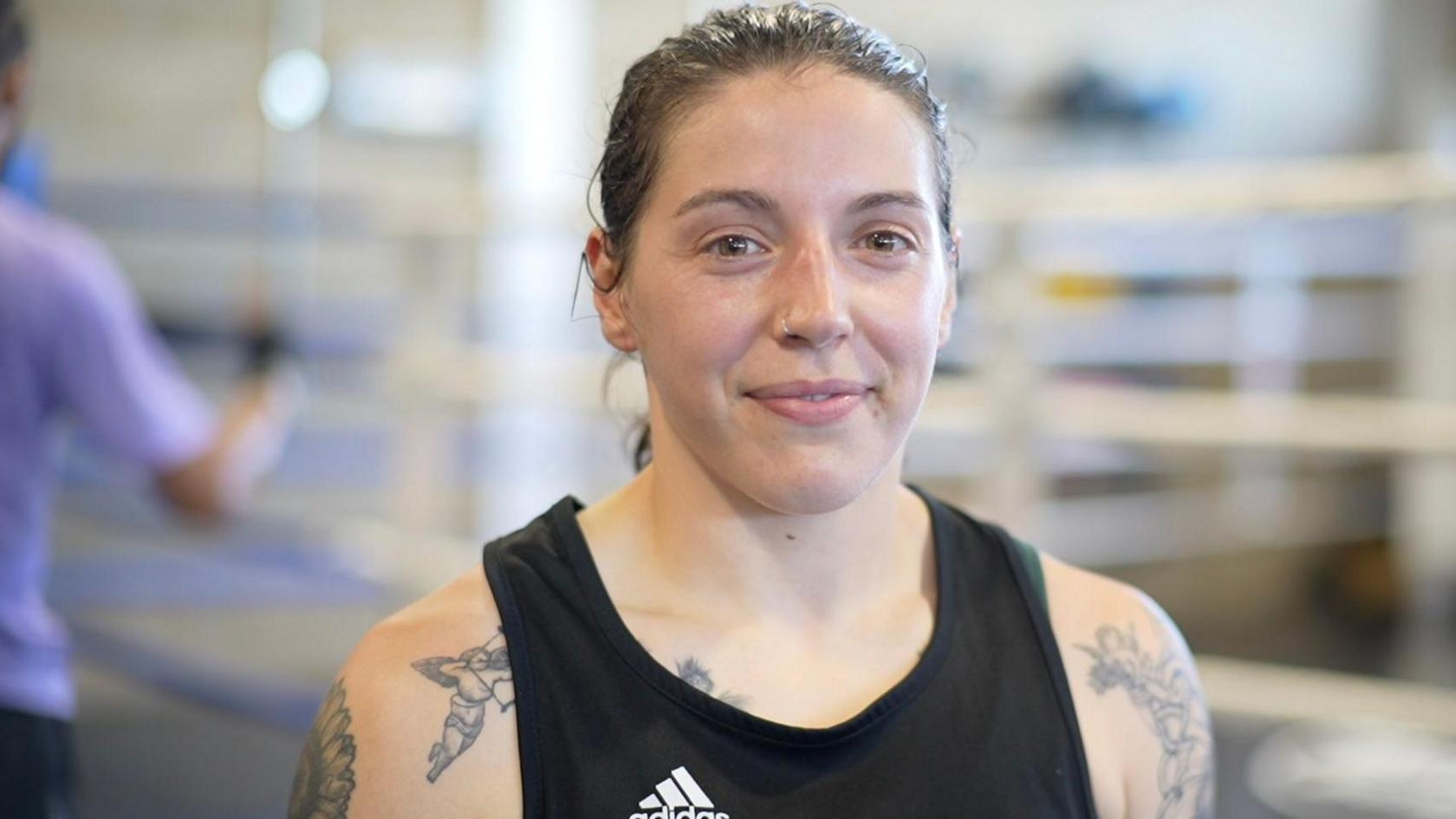 Eilish Tierney smiles at the camera. She wears a black adidas vest and has tattoos on both arms and shoulders - the visible ones are of Cupid flying with an arrow. Her brown hair is tied back and she has a silver nose ring.
