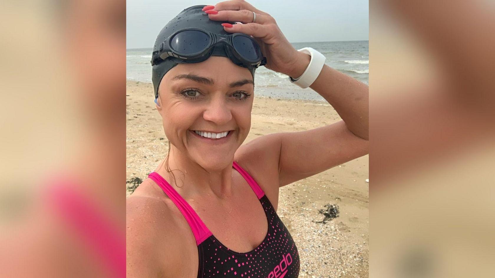 Rebekah Kefford looks at the camera and smiles in a selfie. She is wearing a swimming costume with pink shoulder straps and a black swimming hat and black goggles on her forehead. She is standing on a beach.