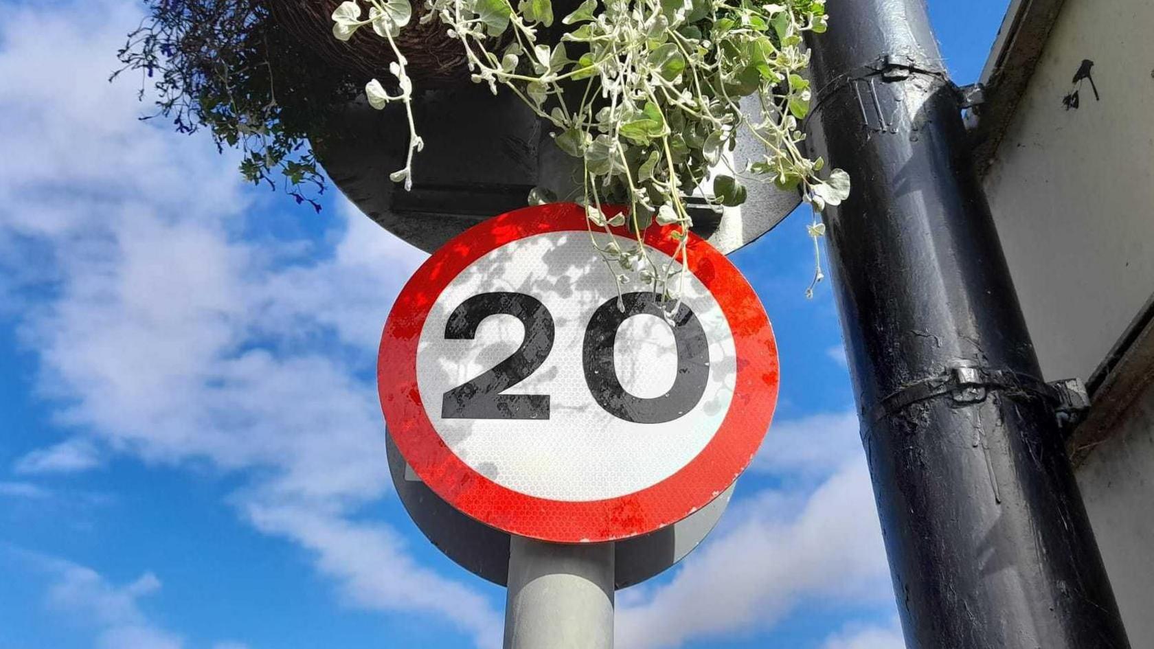 Circular speed limit road sign with 20 written on it on a white background with a red border, below a hanging basket