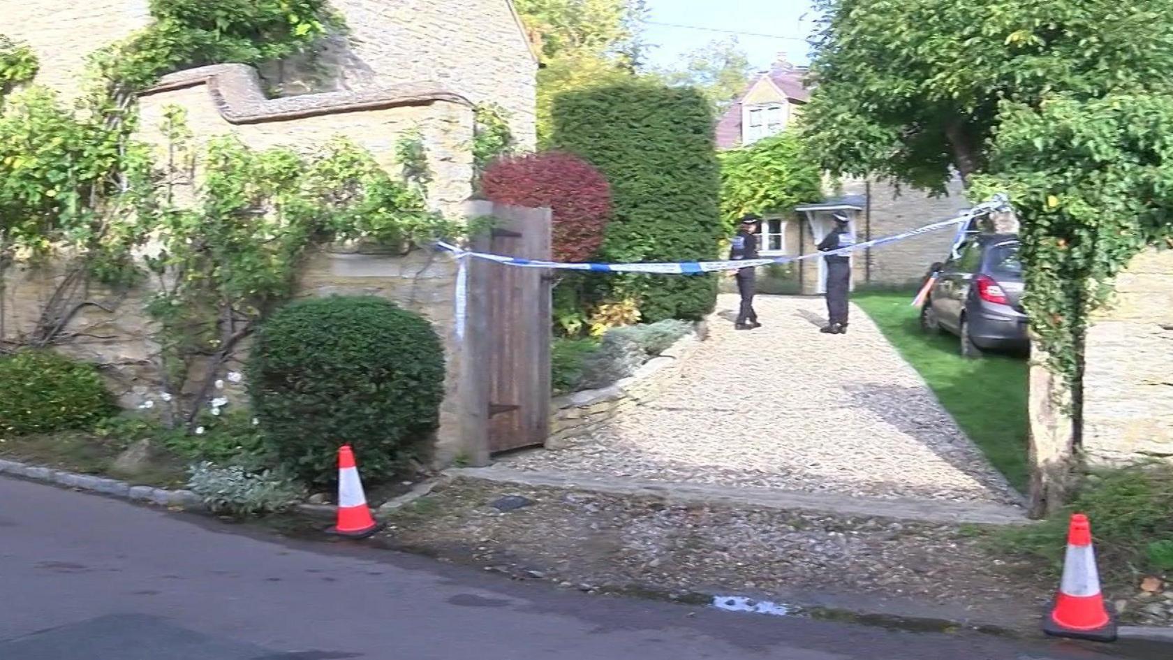 A stone driveway off a road with cones in place at the entrance - it leads to a house where two police officers are standing near the front door.