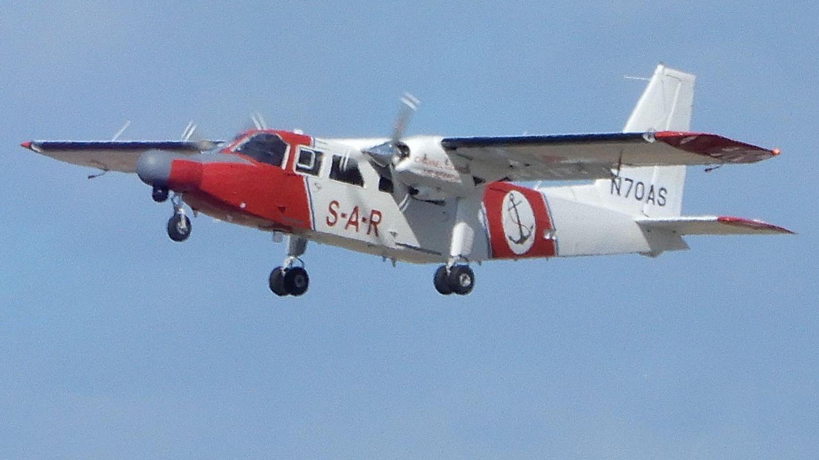 The red and white Channel Islands Air Search plane flying in daytime. It has SAR printed on its side and a logo incorporating an anchor. On its tail fin there is number N70AS. Its wheel are down.