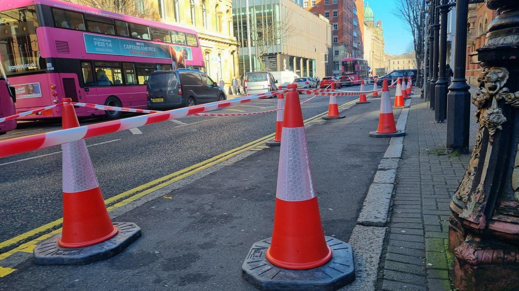 Rows of fluorescent orange traffic cones with striped tape strung between them on a footpath along Bedford Street in Belfast