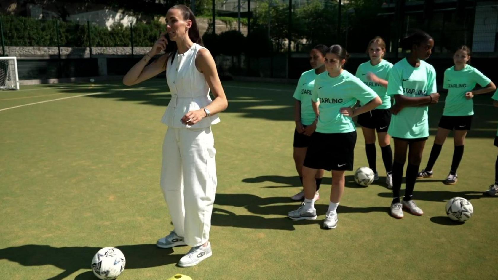 A group of girls in green football kit watch as Jill Scott, dressed in a white sleeveless top and white trousers, demonstrates her football skills. 