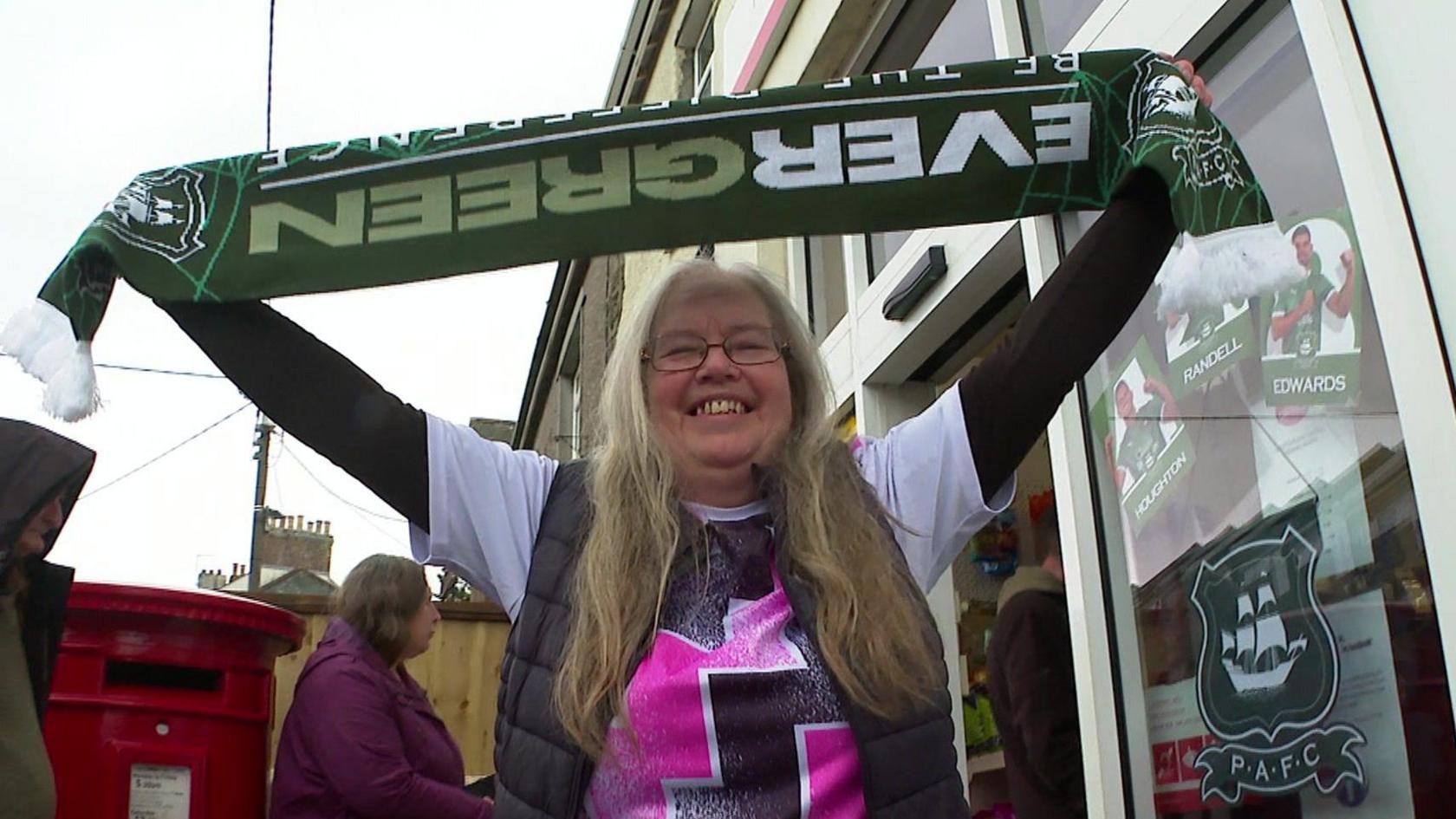 A smiling woman with long hair and glasses holds up a green and white Plymouth Argyle football scarf outside a post office. She is wearing a black thin puffer over a pink and white top Argyle top with a long sleeve black top under that. Behind her, other people are visible, along with a red postbox and posters featuring Plymouth Argyle players on the shop window.