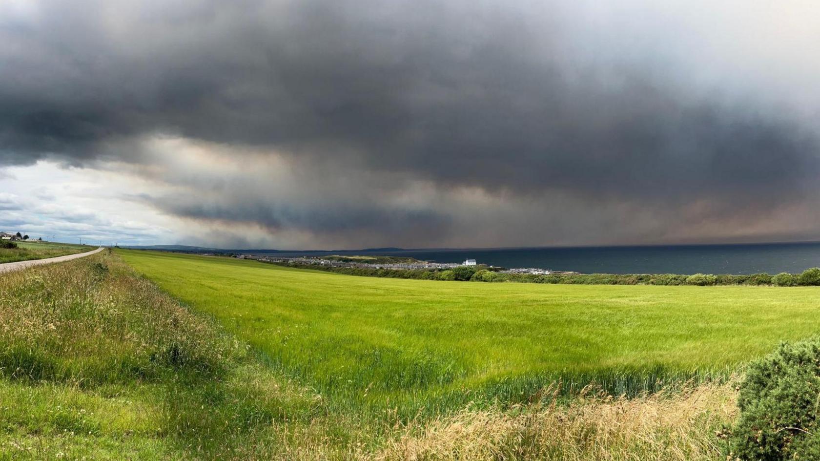 Thick grey smoke fills the sky above the Moray Firth. In the foreground is a grassy field and a road.