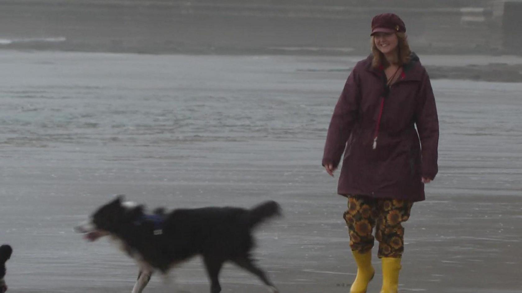 A woman wearing a hat, coat and welly boats with a Collie dog on the beach