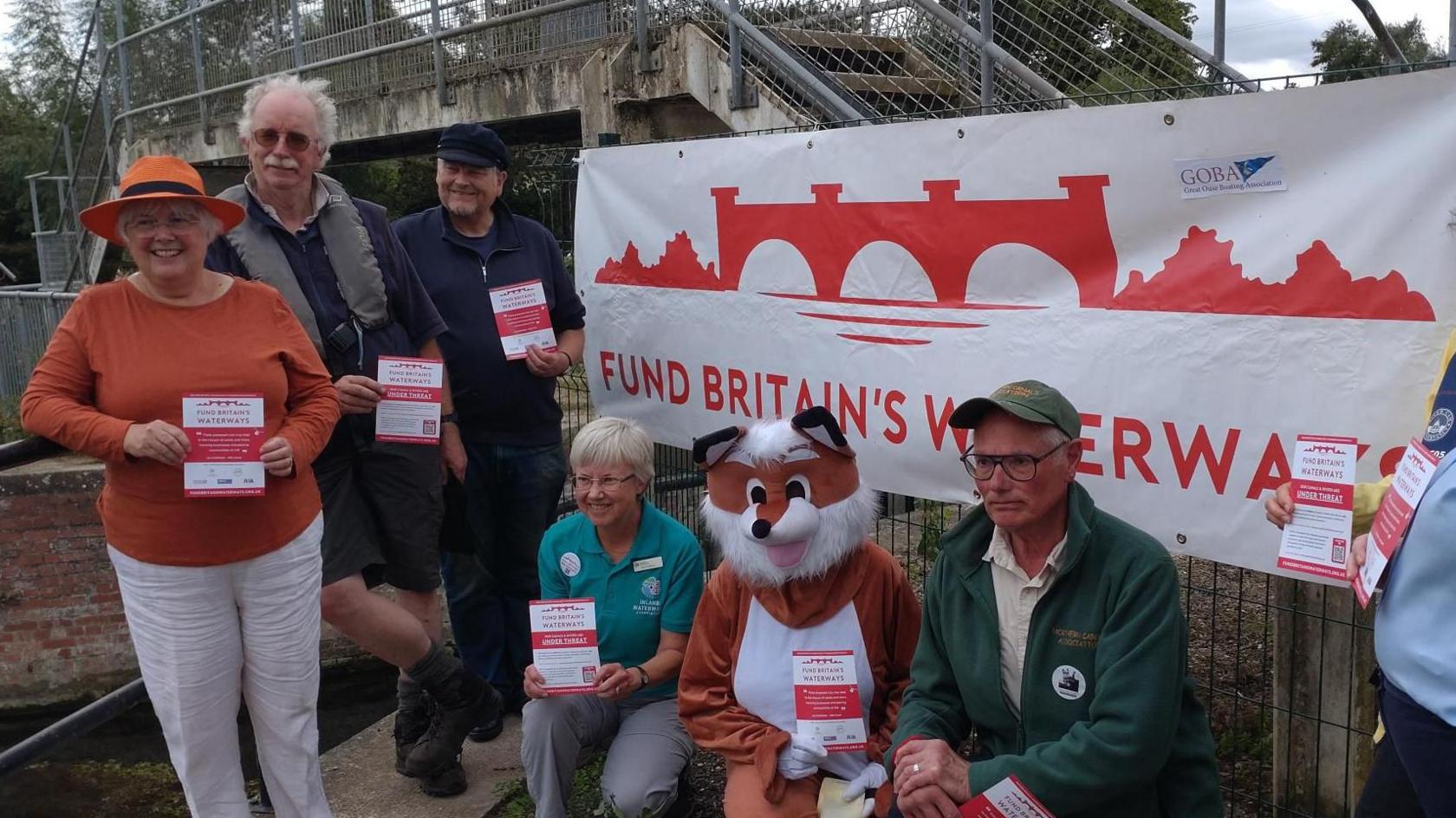 A white poster with red writing says 'Fund British Waterways.' A foot bridge over a river is behind it. A group of people, including one dressed in a full fox outfit hold leaflets.