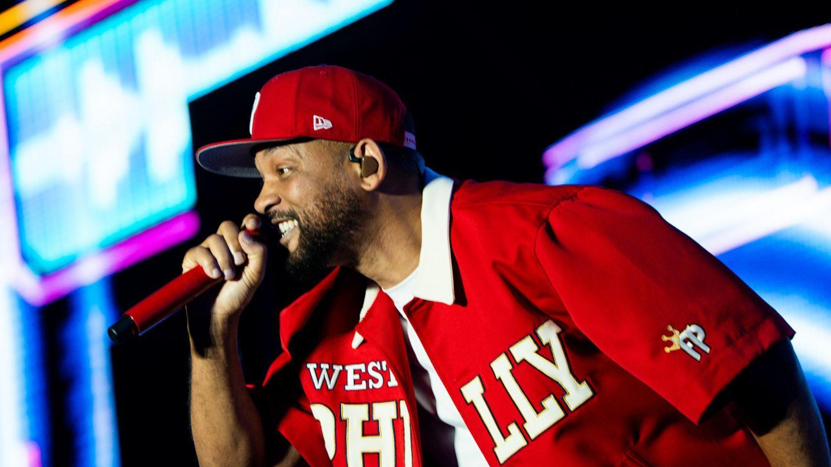 Will Smith sings into a red hand-held microphone. He is wearing a red cap and a red shirt which is open. 'West Philly' is written across the shirt in white. He is on a brightly-lit stage.