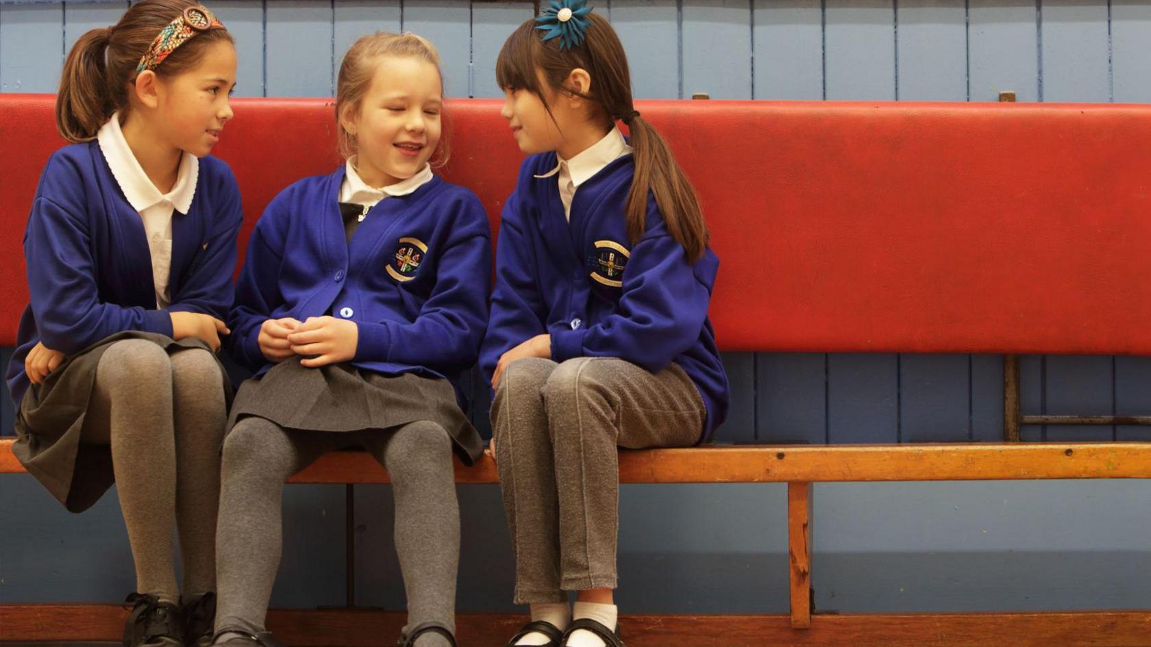 Three girls at primary school wearing blue and grey uniforms. Two are wearing skirts whilst one is in a pair of trousers. 