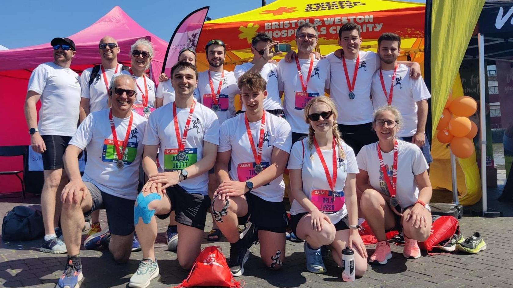 Fourteen people wearing red Bristol Half Marathon medals and white t shirts supporting Bristol & Weston Hospitals Charity, shorts and trainers.