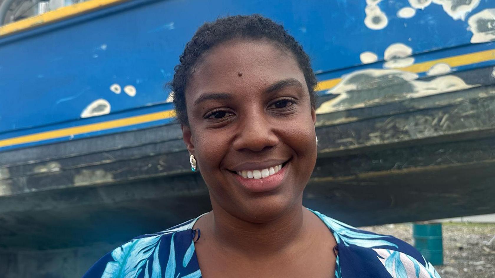 Dr Shelly Ann Cox, short-haired and smiling, wearing a two-tone blue blouse and standing in front of the weather-beaten hull of a boat