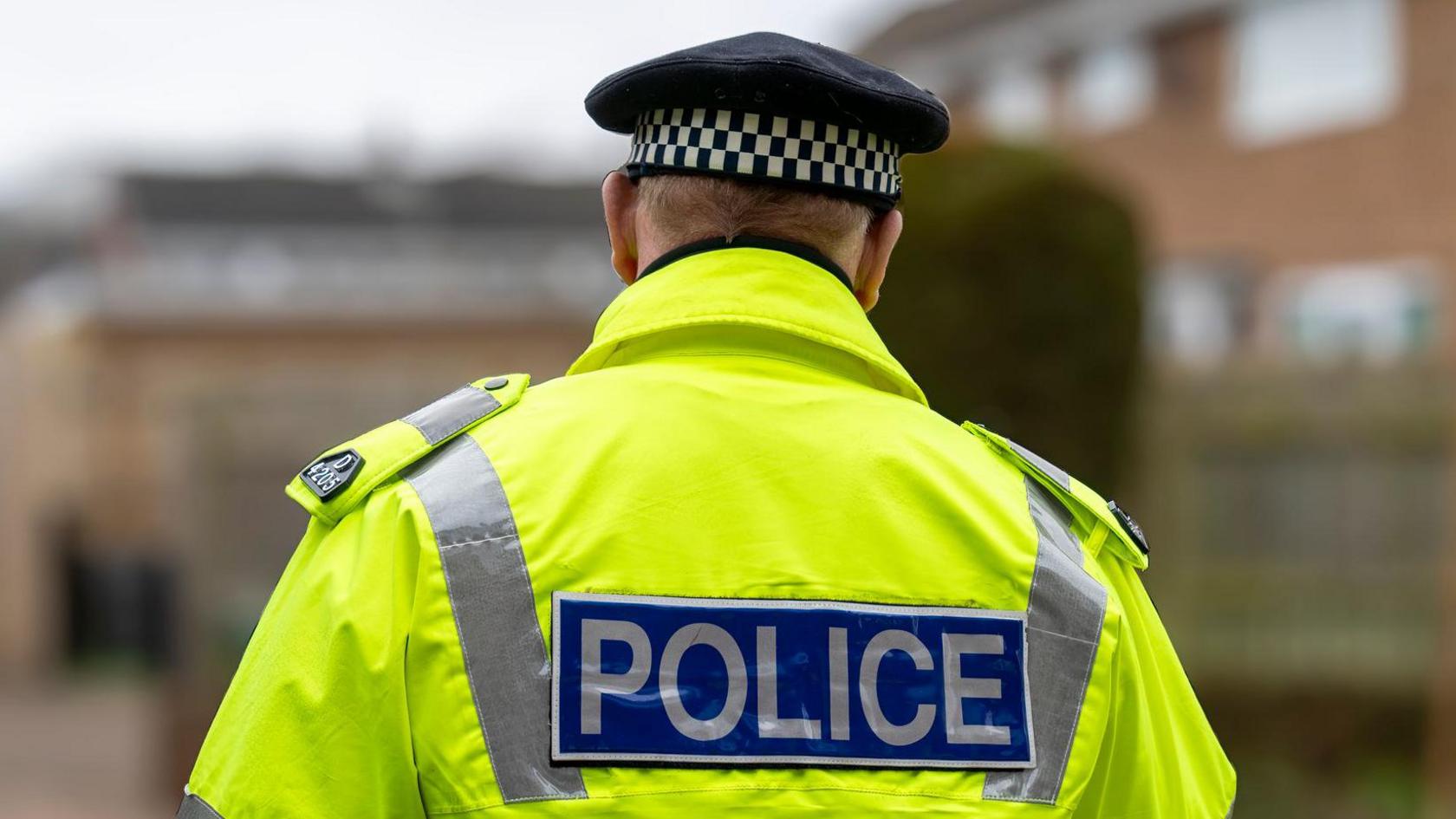 Close-up of a police officer from behind. He is wearing a yellow hi-vis jacket with the word 'POLICE' written in silver writing on a blue background. The road in front of him is out of focus