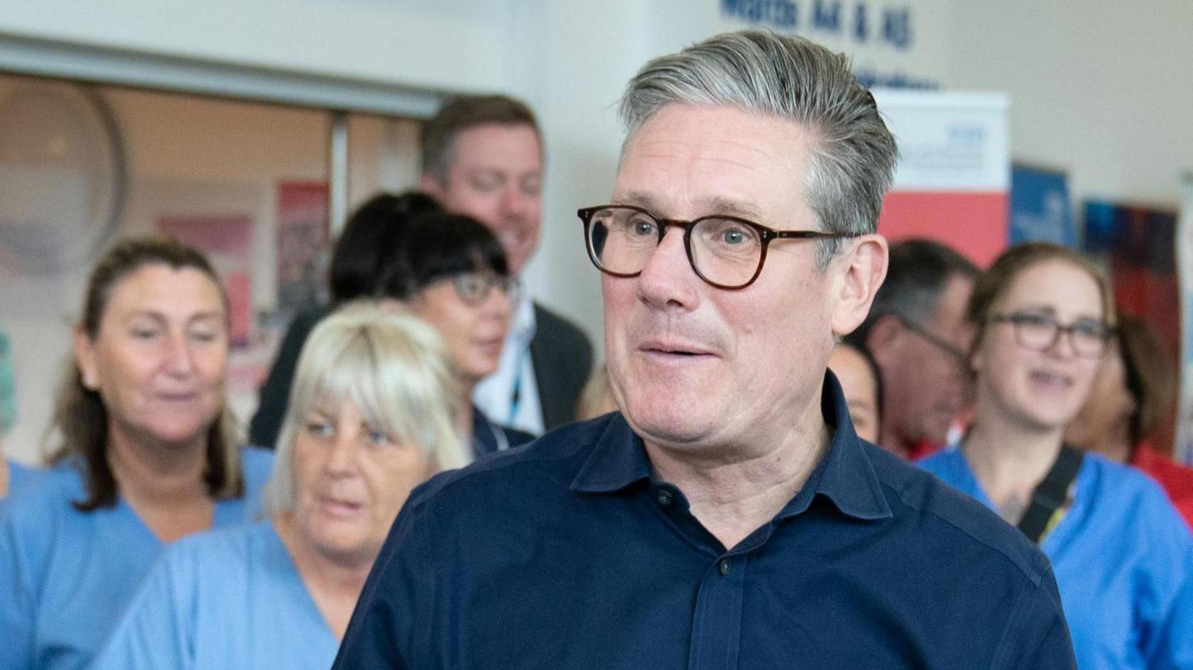 Sir Keir Starmer, in a blue open-necked shirt and dark rimmed glasses, standing in front of a group of women in blue hospital worker shirts