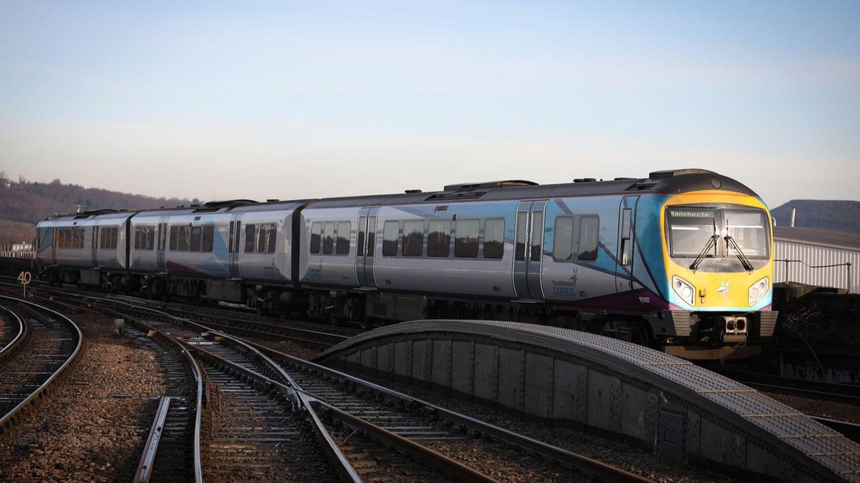 A silver and blue TransPennine Express train on tracks. The front of the train is yellow and features a small sign in the left corner that reads "Manchester".