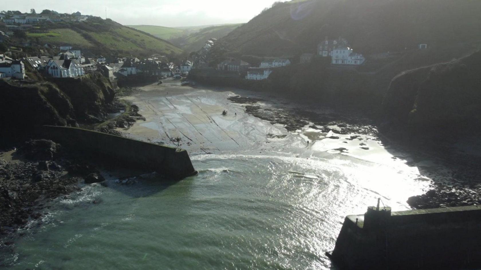 A drone shot of Port Isaac, there are two piers protecting the harbour, houses line the cliffs above the village.