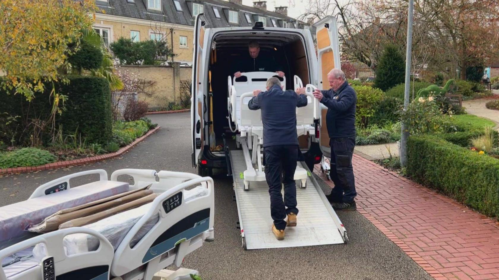 Three men unload a cuddle bed down a ramp from a van, with another cuddle bed already unloaded.