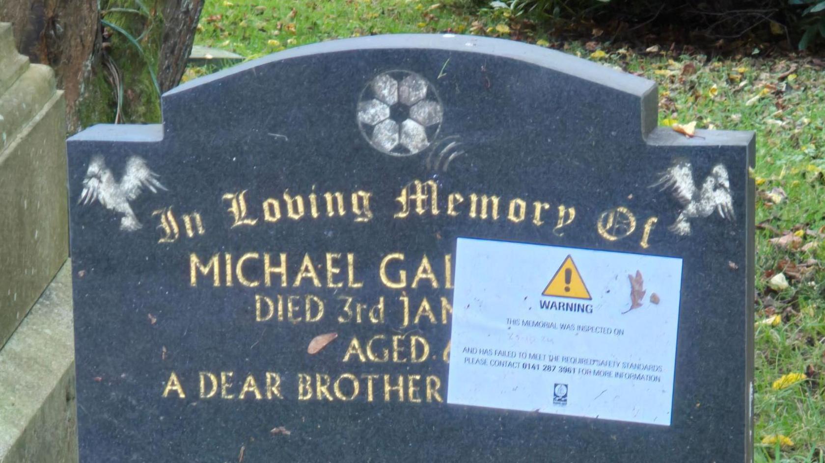 A  black gravestone with gold lettering at Riddrie Park Cemetery - the first name of the deceased is Michael, but the surname has been covered by a large sticker saying WARNING with a exclamation mark in a yellow box   