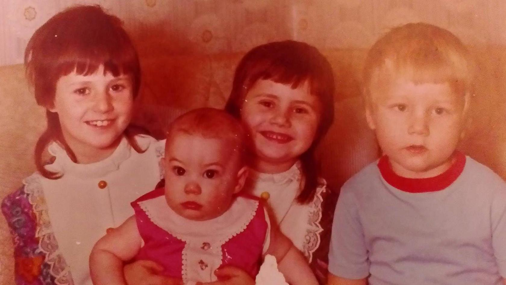 Vintage image of a group of young children holding a baby. The two girls are wearing 1970s style dresses with high lace collars, and have dark uneven fringes and mullets, the young blonde-haired boy is wearing a pale blue t-shirt with a red collar and the baby girl is wearing a red sleeveless dress with buttons.