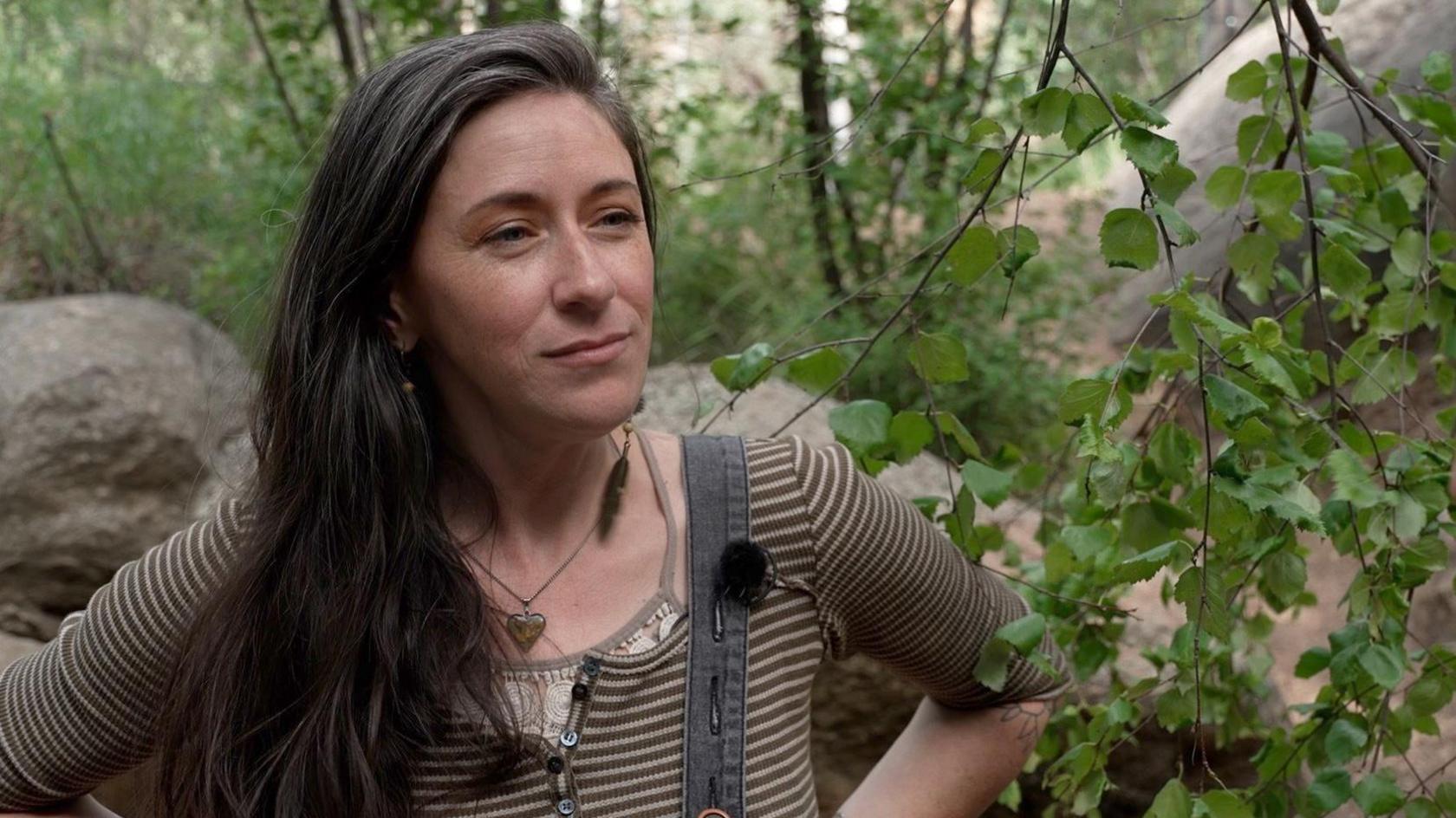 Wild Mother, real name Desiree, a young woman with long brown hair and wearing dungarees, standing amid trees and rocks in the mountainous part of Colorado where she lives