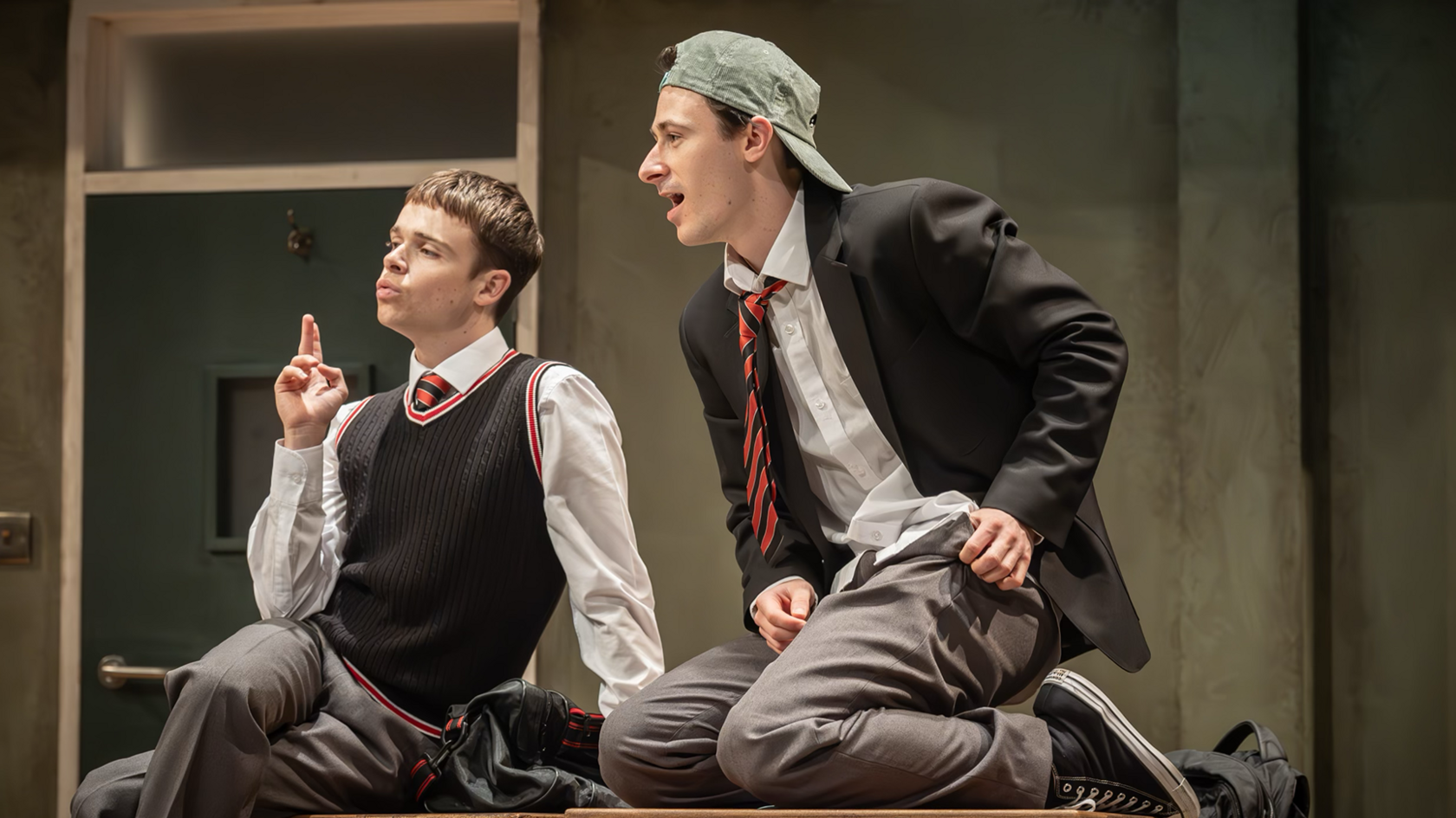 Two boys sit on a table, wearing school uniform. They are looking out to the left of the photo. The are clearly on a stage.