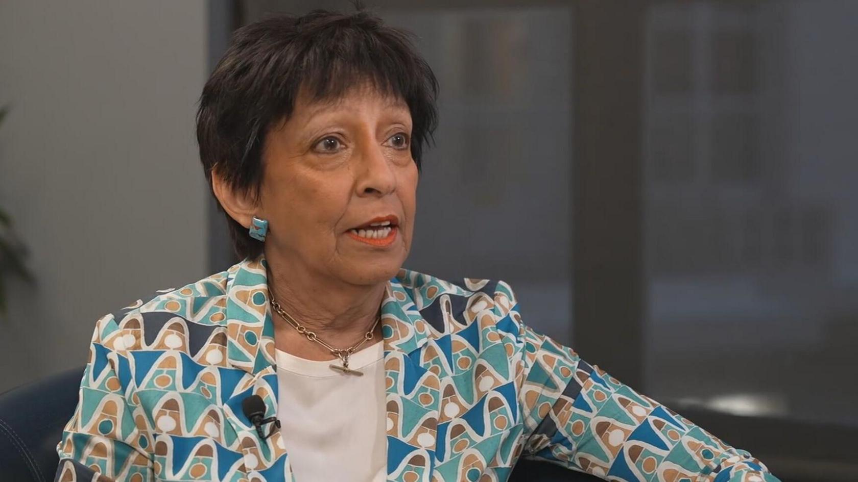 A woman with black hair speaks while seated. She is wearing a patterned blue, white and brown jacket over a white top, with a window and a white wall behind her.