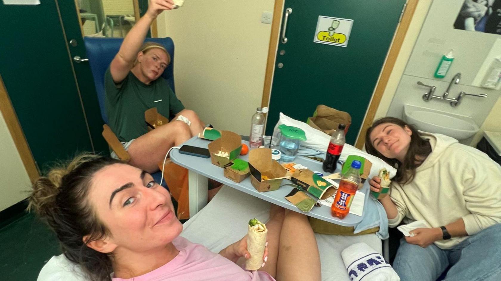 Emma Wassell in her hospital bed, eating food from the table in front of her while two of her teammates smile for the camera, also with food in hand