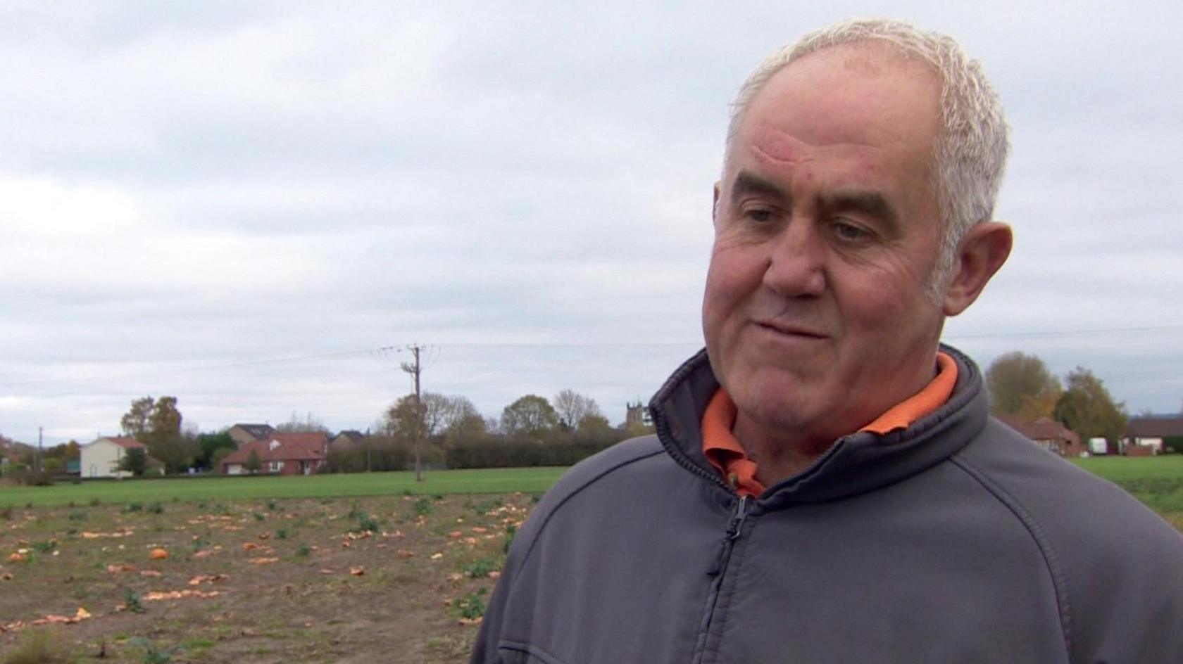 An image captured of Patrick Wilson, who has short grey hair and wears a grey zip-up jacket and orange polo shirt, standing in his pumpkin patch. 