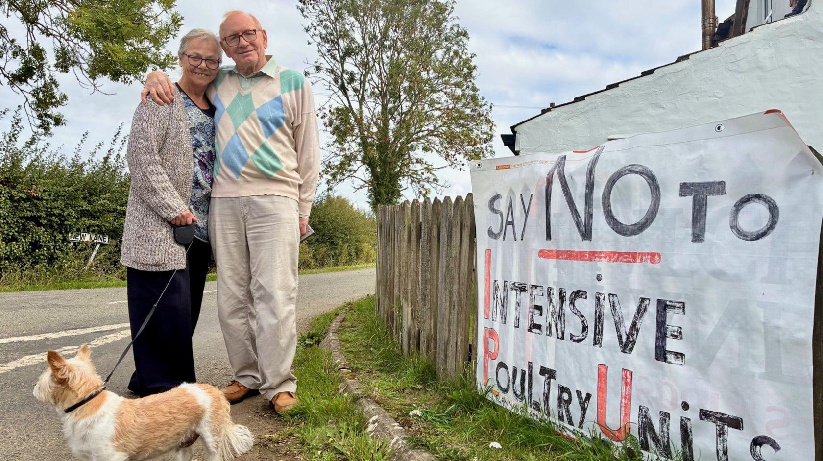 Maggie and Lyndon Brace stand outside of their home in Toynton St Peter in Lincolnshire. Beside them is a banner attached to a hedge expressing opposition to a nearby poultry unit proposal.