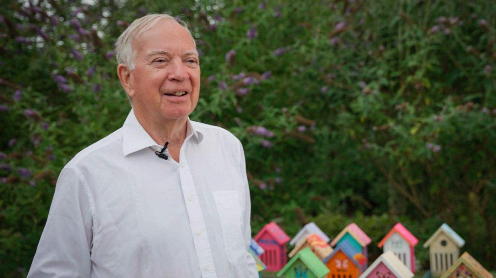 Hugh Ellerton, who has white hair and wears a plain white shirt, faces the camera with the greenery of a garden behind him and brightly coloured miniature houses designed for butterflies shown on the ground.
