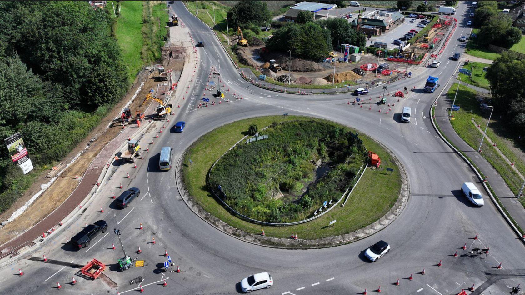 An aerial view of the roundabout shows cones and workers at all four junctions and cars travelling round in a clockwise direction. There is a grass island in the middle of the roundabout.