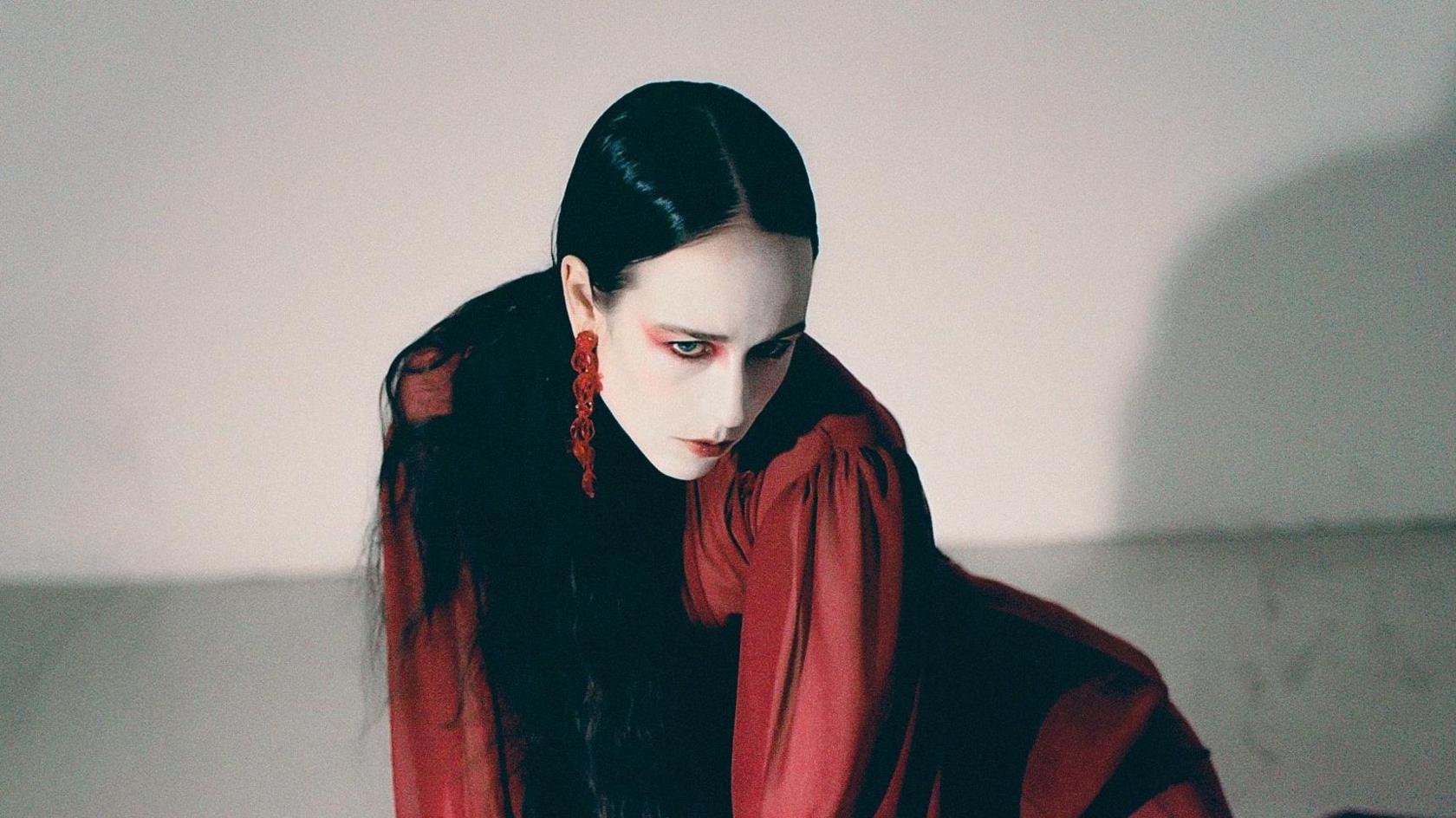 Allie X posing in a white room. She is wearing a red dress and is staring at the ground.