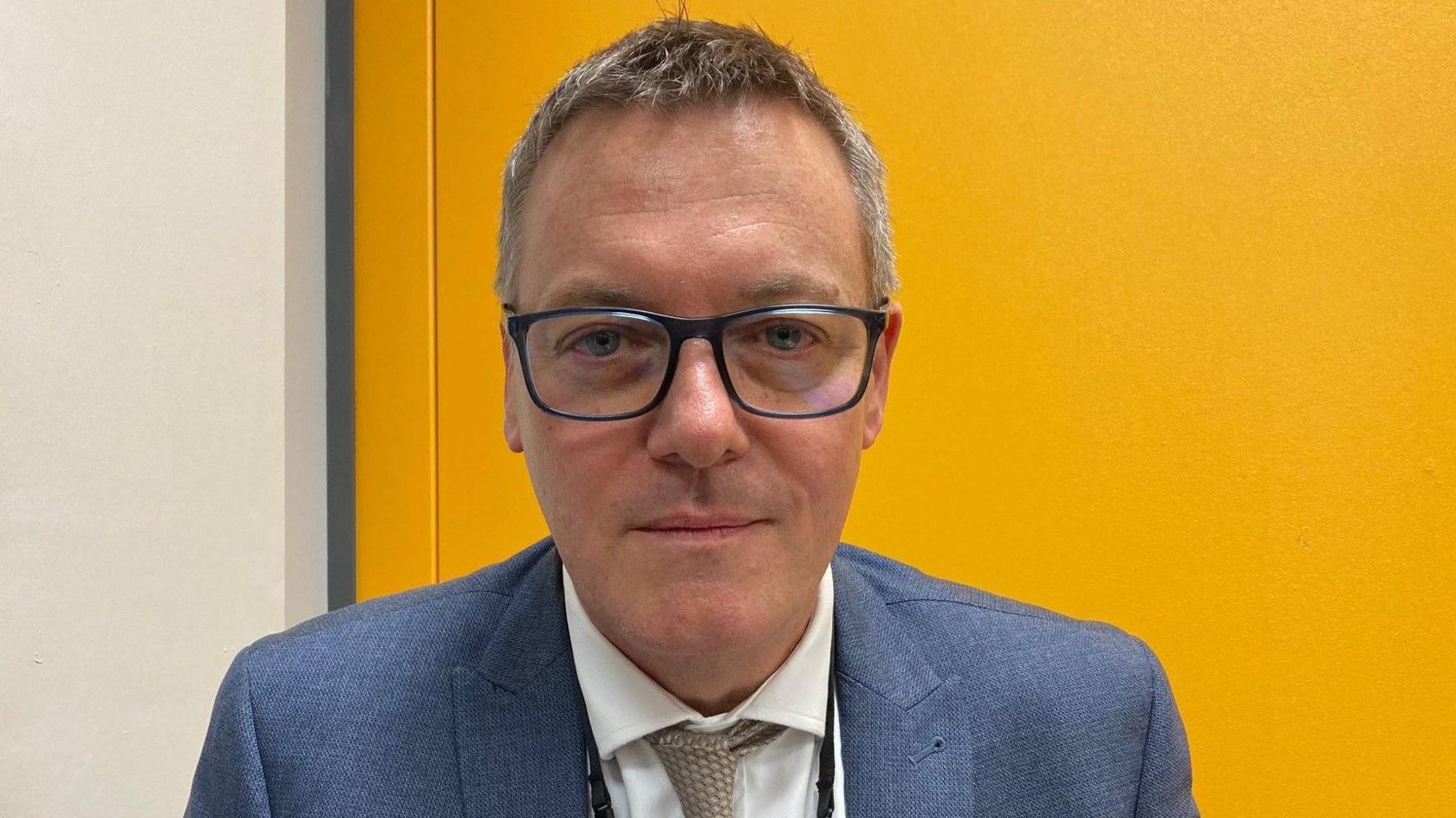 A man wearing glasses, a shirt and tie and blue jacket. 