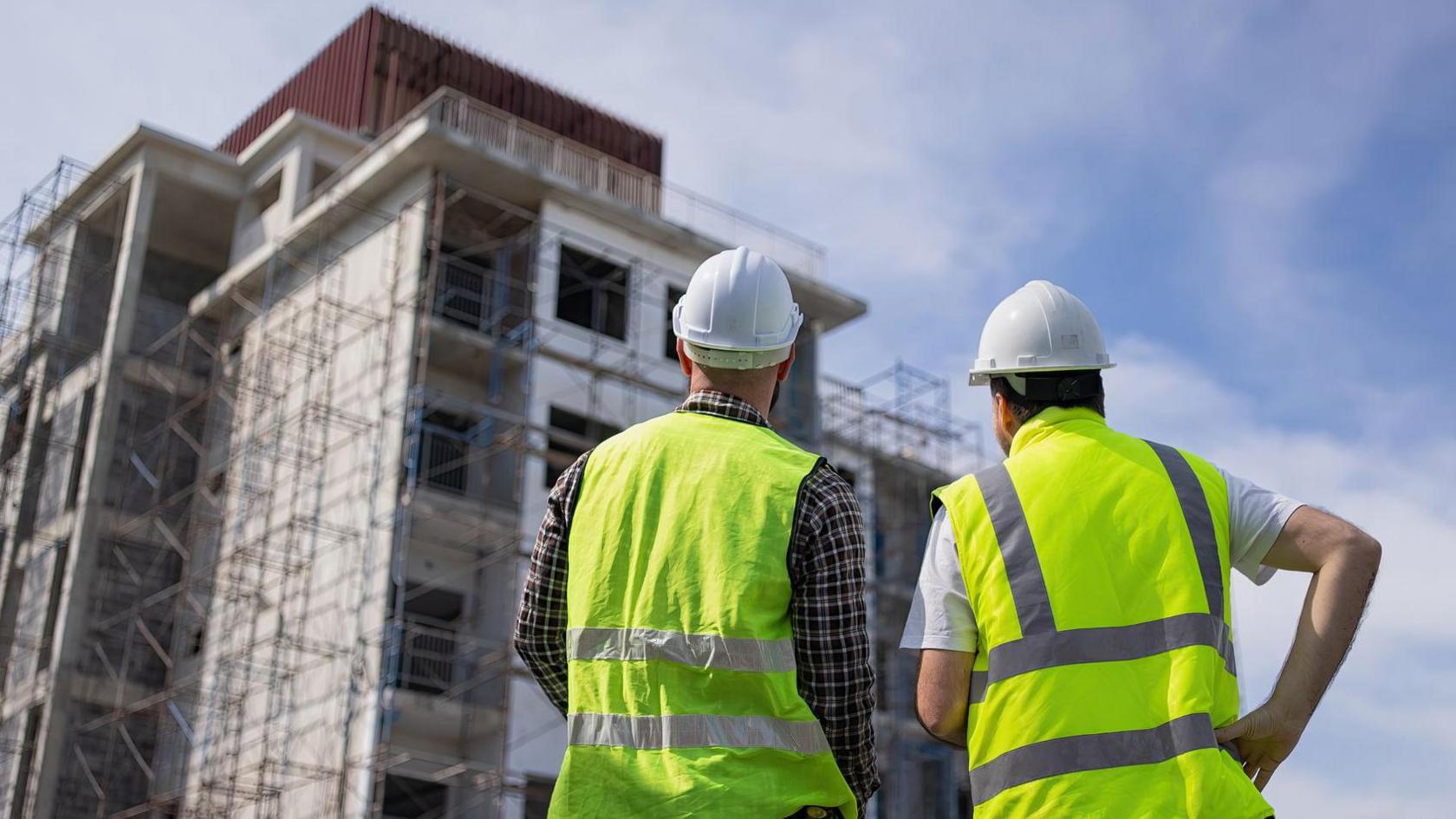 Two men in hi-vis stand next to each other and look up to a  multi-storey residential building being constructed in front of them. The man on the left has a checked shirt, hi-vis and a white hard hat on. The man on the right has a white t-shirt on with his hi-vis and white hard hat.