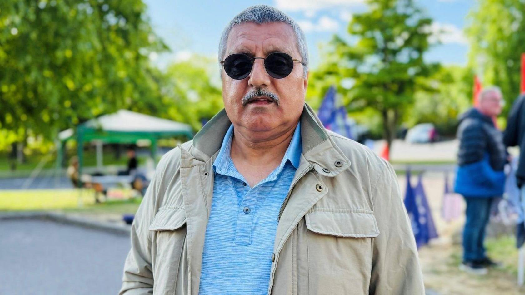 Man - Saddiq Ahmed - looking at camera, he has greying hair, is wearing dark round sunglasses, and light blue top which is unbuttoned at the top, and a beige jacket.