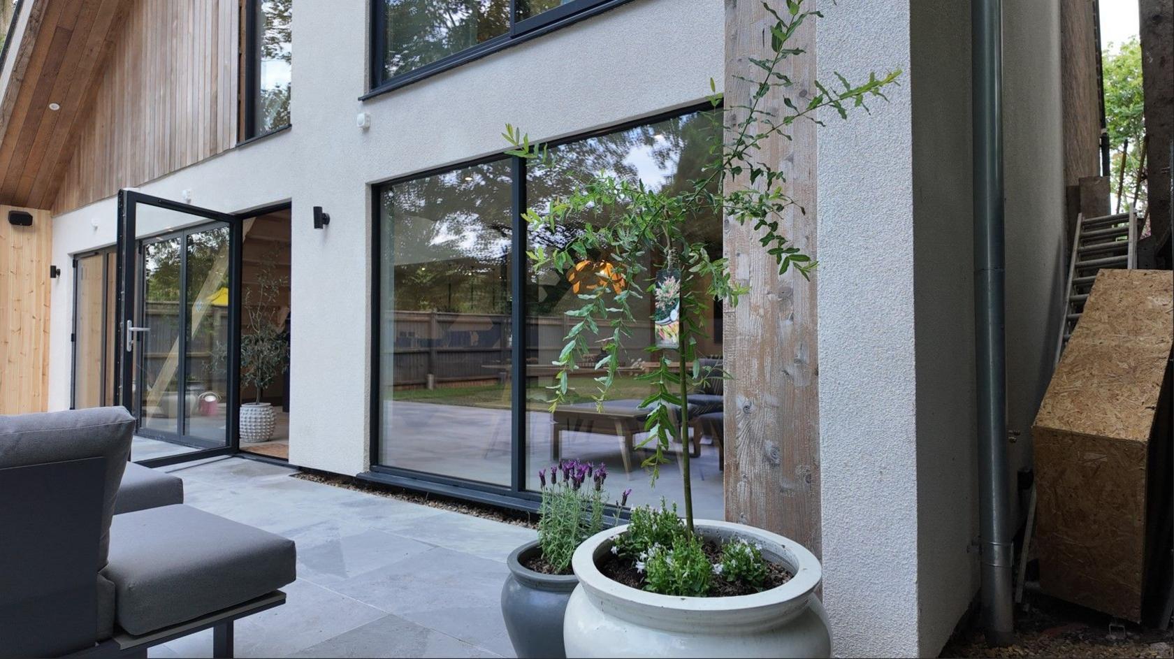 Big glass doors and windows at the back of a refurbished house give way to a patio area with chairs and potted plants.