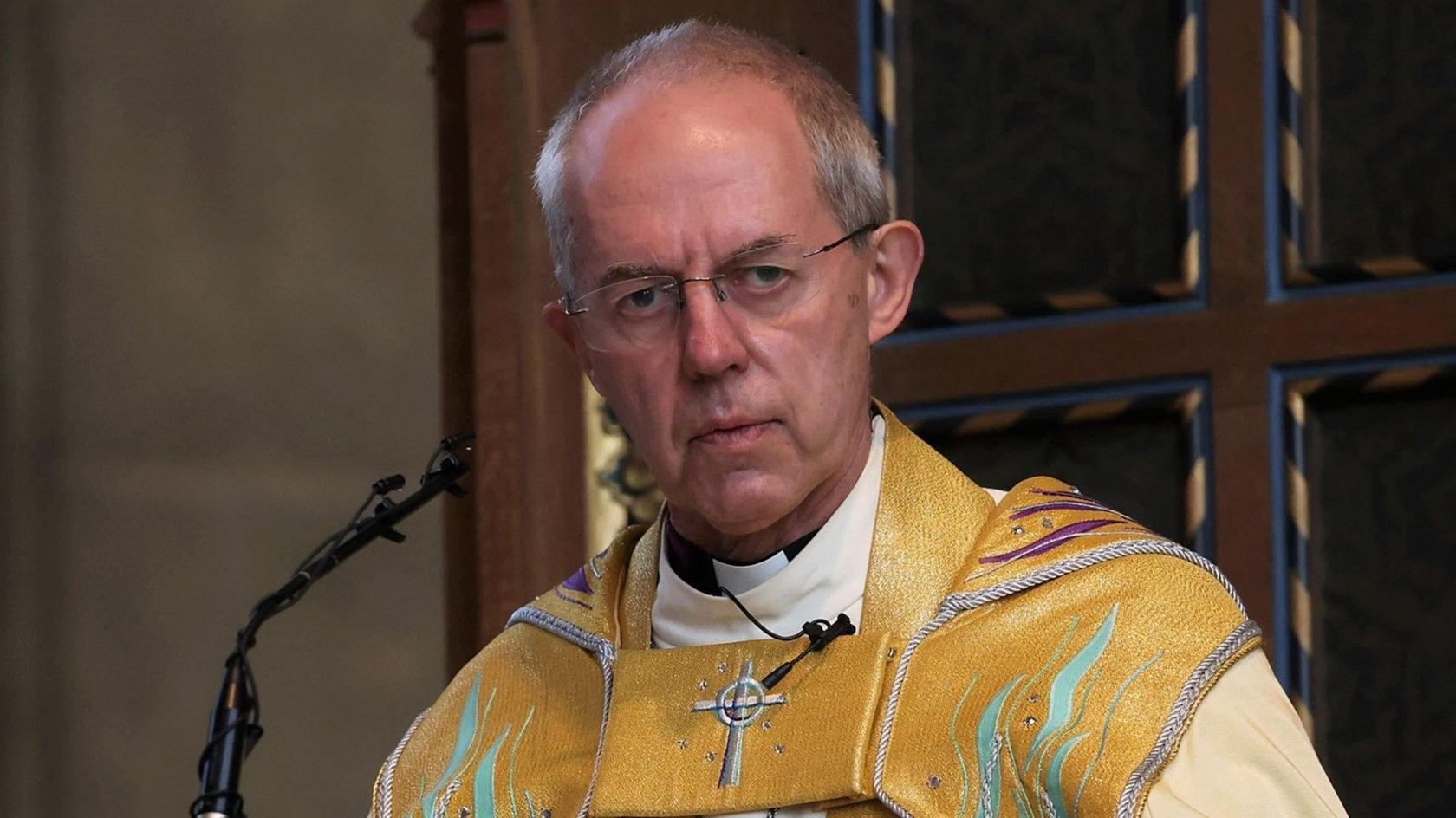 Archbishop Justin Welby at a pulpit in yellow robes. He is wearing glasses, has short grey hair and is looking at the camera with a serious expression.