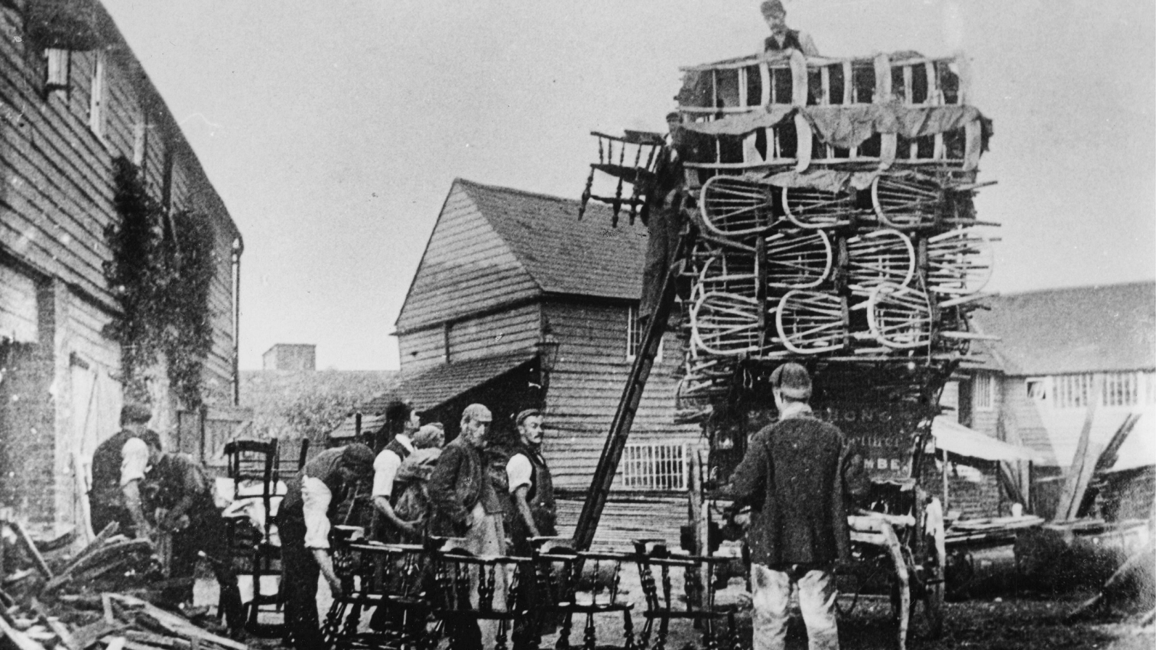 A black and white photo showing chairs being loaded onto a wagon by a group of men in caps, at Gibbons chair factory, off Oxford Road, High Wycombe in about 1895. 