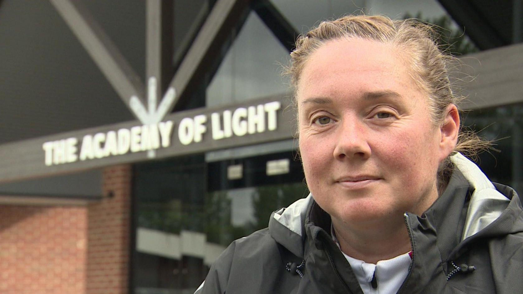 Mel Reay sits in front of the main entrance to the Academy of Light in South Tyneside, she is dressed in a Sunderland AFC tracksuit which is black and grey with a white hoodie.