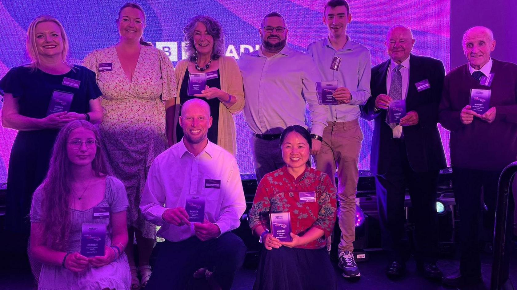 The winners photographed in a group at the ceremony, all smiling and smartly dressed holding their glass awards. Three of them are kneeling in front of a row of winners. They are standing in front of a screen which is purple and has the white BBC Radio Cornwall logo on it.