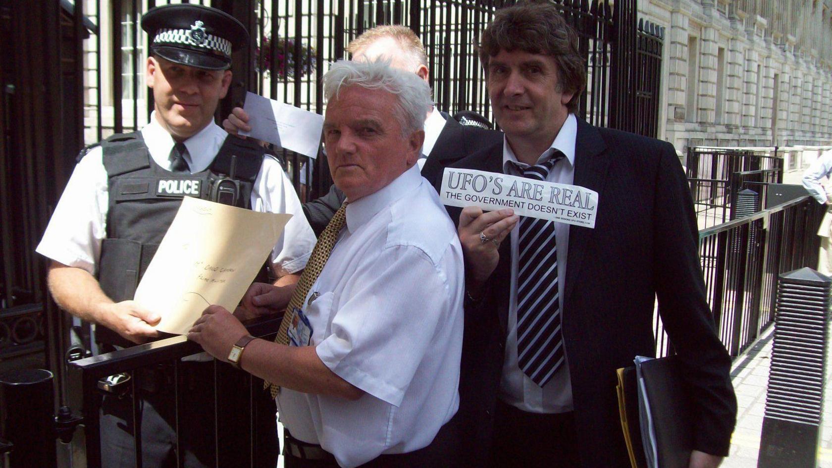 Outside the gates to Downing Street a man with white hair and wearing a white shirt with yellow tie is handing an envelope to a male police officer. To the man's right is a man in dark suit, white shirt and blue and white striped tie. He is holding up a sign saying "UFOs are real".