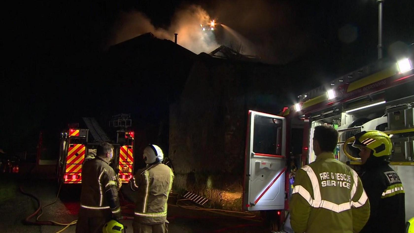 Four firefighters next to two appliances with smoke from a fire in the background at night.