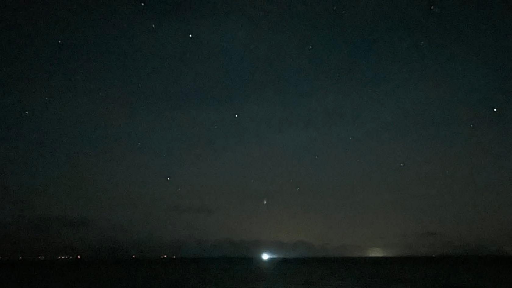 A dark stretch of sky with various bright white dots scattered across it. There is a light on the horizon.