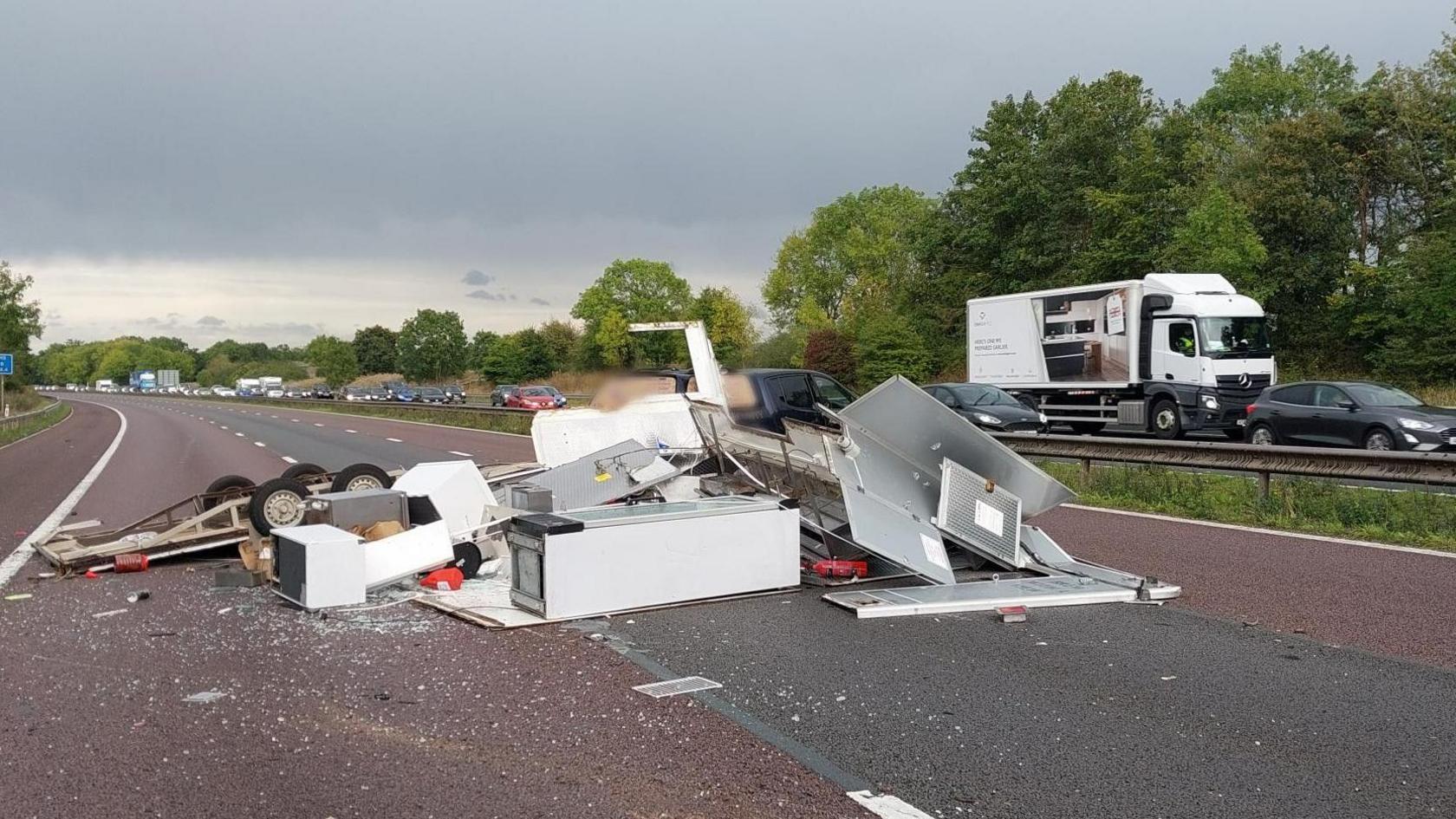 White goods, large building materials and broken glass can be seen scattered across the carriageway. The road in front of it is completely clear due to the blocked traffic.