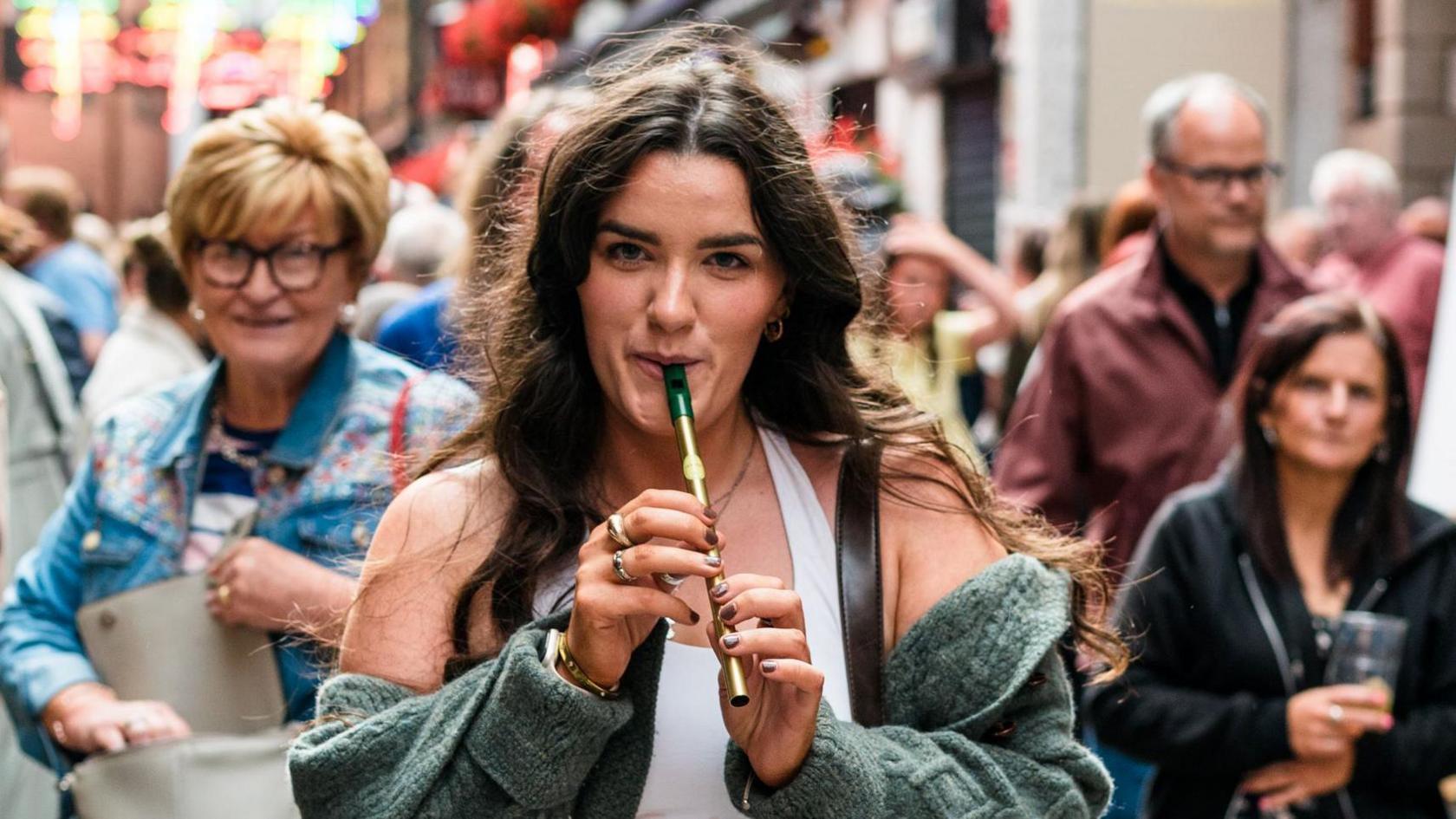 A woman with long brown hair, playing a tin whistle. There is a crowd of people behind her. 
