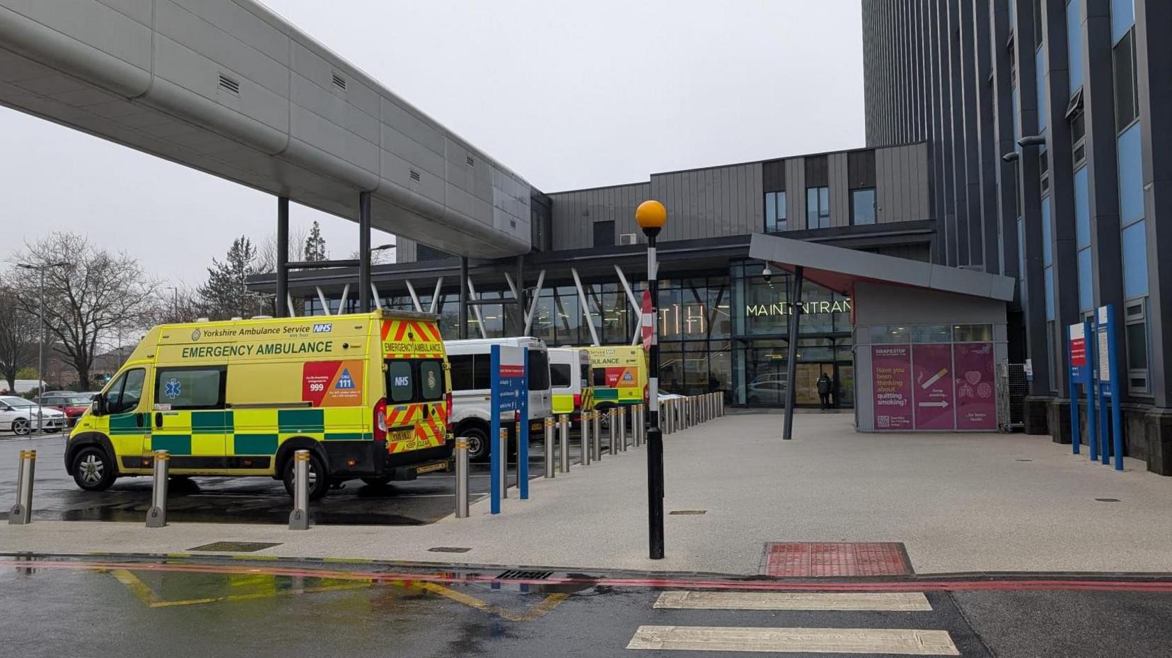 Yellow and white ambulances parked outside the concourse of a large hospital building with a grey and blue tower block to the right, a modern glass-fronted atrium in the centre, and an enclosed grey walkway overhead.