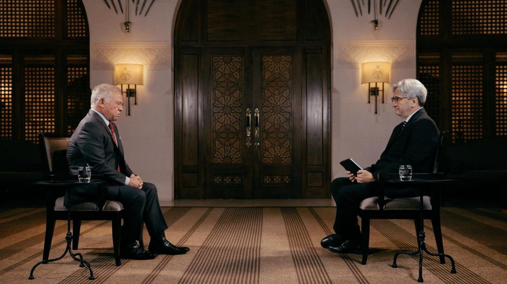 King Abdullah sitting in a chair opposite Fergal Keane, also on a chair. The two men are about a metre apart, and next to them are tables with a glass of water on. Behind them are ornately-patterned, wooden double-doors and two wall lamps.