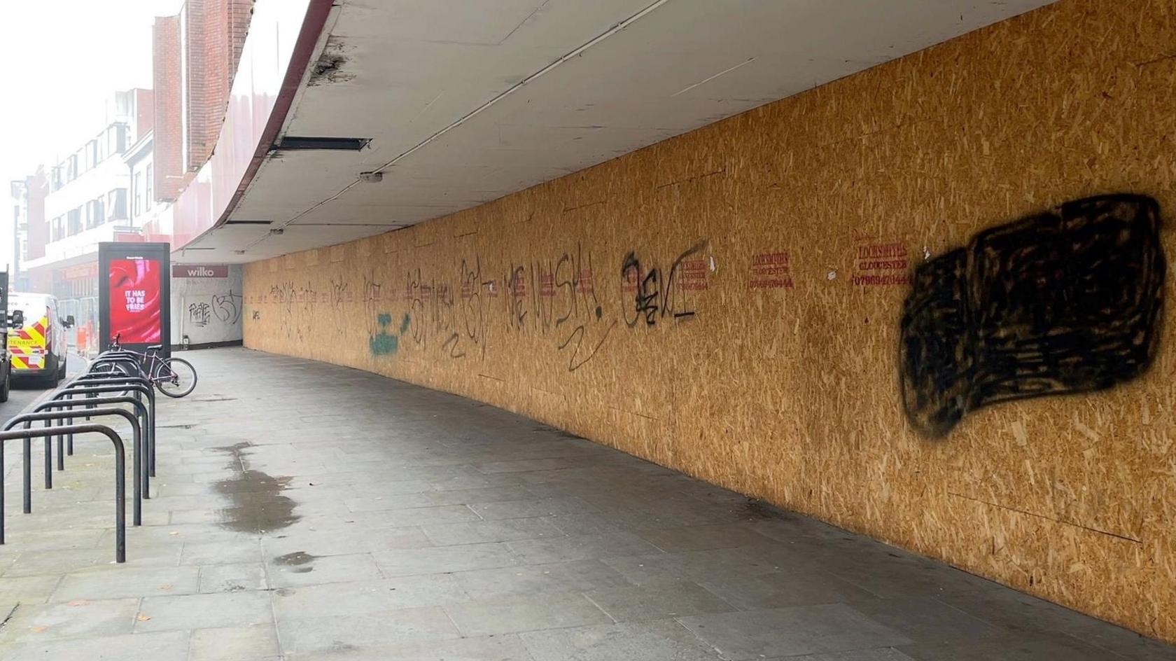 This image shows a boarded up shop front, looking down the street. The willkos has a long shop front stretching down the street. On the wooden boards, sitting on the right hand side of the image is graffiti. On the right hand site is some bike racks and in the distance a highway maintenance van. 